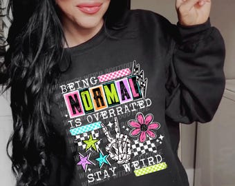 Può includere: Una felpa nera a girocollo con una stampa grafica che riporta il testo "BEING NORMAL IS OVERRATED STAY WEIRD". Il design include una mano di scheletro che fa il segno di pace, un fiore rosa, stelle colorate e motivi a scacchi.