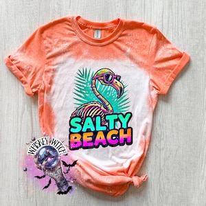 May include: Orange and white t-shirt with a flamingo graphic wearing sunglasses and the words "Salty Beach". The flamingo is in front of green palm leaves. The shirt has a tie-dye effect.