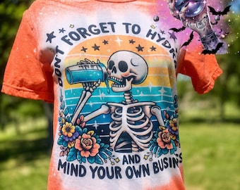May include: Orange short-sleeved t-shirt with a skeleton graphic drinking from a blue container. The shirt has the text "DON'T FORGET TO HYDRATE AND MIND YOUR OWN BUSIN'S" in a colorful design. The shirt has a bleached effect.