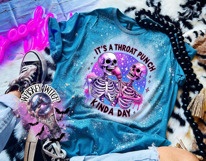 May include: A turquoise blue t-shirt with a bleach-splattered effect. The shirt features a graphic of two skeletons boxing with the text "It's a throat punch kinda day".