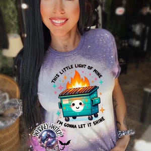 Purple  t-shirt with a bleach-splattered design featuring a cartoon dumpster on fire with the text "This little light of mine I'm gonna let it shine".