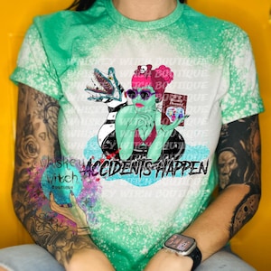 May include: A green, white, and black bleached t-shirt with a graphic of a woman with pink hair holding a book that says "Recently Deceased" and a creature from the black lagoon. The text "Accidents Happen" is printed below the graphic.