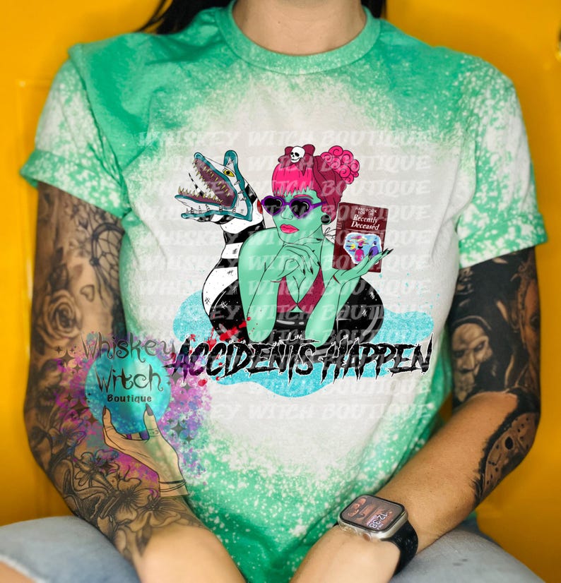 May include: A green, white, and black bleached t-shirt with a graphic of a woman with pink hair holding a book that says "Recently Deceased" and a creature from the black lagoon. The text "Accidents Happen" is printed below the graphic.