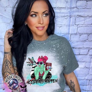 May include: A gray t-shirt with a bleached effect featuring a green cartoon witch holding a book that says "Friends Forever" and a black and white striped creature. The text "Accidents Happen" is printed below the witch.