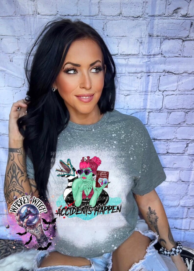 May include: A gray t-shirt with a bleached effect featuring a green cartoon witch holding a book that says "Friends Forever" and a black and white striped creature. The text "Accidents Happen" is printed below the witch.