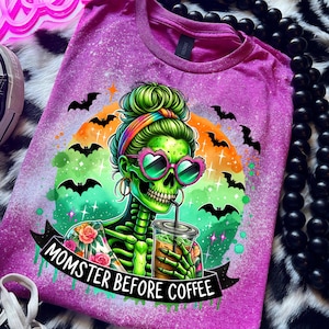May include: A purple t-shirt with a graphic of a green skeleton wearing sunglasses and drinking coffee. The text "MOMSTER BEFORE COFFEE" is on a banner below the skeleton. Bats and a colorful sunset are in the background.