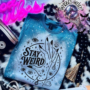 Handmade bleached styles shirt. Close-up of witchy Stay Weird shirt design with celestial artwork, mystical hands, stars, and bold black text.