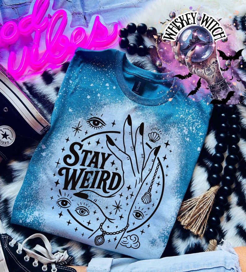 Handmade bleached styles shirt. Close-up of witchy Stay Weird shirt design with celestial artwork, mystical hands, stars, and bold black text.