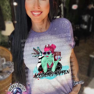 May include: A purple tie-dye t-shirt with a graphic of a green-skinned woman with pink hair wearing sunglasses and holding a book that says "Freddy's Dead". The woman is sitting in a pool with a black and white striped creature. The text "Accidents Happen" is printed below the graphic.