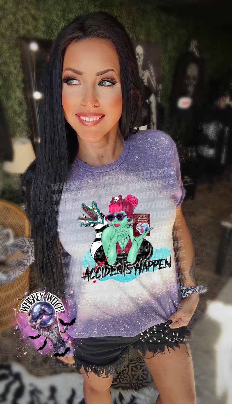 May include: A purple tie-dye t-shirt with a graphic of a green-skinned woman with pink hair wearing sunglasses and holding a book that says "Freddy's Dead". The woman is sitting in a pool with a black and white striped creature. The text "Accidents Happen" is printed below the graphic.