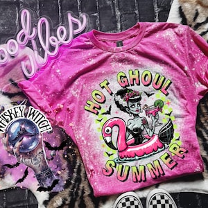 A pink and bleach splattered effect  t-shirt with a graphic of a zombie woman in a flamingo pool float, holding a cocktail. The text "Hot Ghoul Summer" is printed on the shirt.