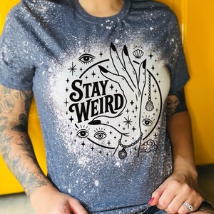 Handmade bleached styles shirt. Close-up of witchy Stay Weird shirt design with celestial artwork, mystical hands, stars, and bold black text
