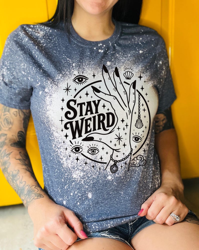 Handmade bleached styles shirt. Close-up of witchy Stay Weird shirt design with celestial artwork, mystical hands, stars, and bold black text