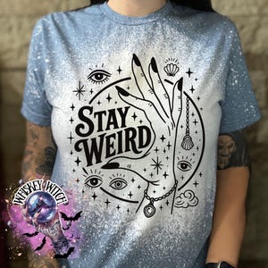 Handmade bleached styles shirt. Close-up of witchy Stay Weird shirt design with celestial artwork, mystical hands, stars, and bold black text