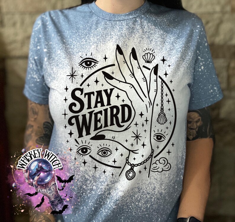 Handmade bleached styles shirt. Close-up of witchy Stay Weird shirt design with celestial artwork, mystical hands, stars, and bold black text