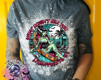 May include: A gray t-shirt with a bleach-splattered design featuring a green-skinned woman in a coffin floating in a pool. The text "What Doesn't Kill You Disappoints Me" is printed in a circle around the woman.