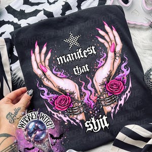 May include: Dark gray t-shirt with a graphic of hands, roses, and the text "manifest that shit." The design includes a checkered star and purple flames. A "Whiskey Witch" sticker is visible.