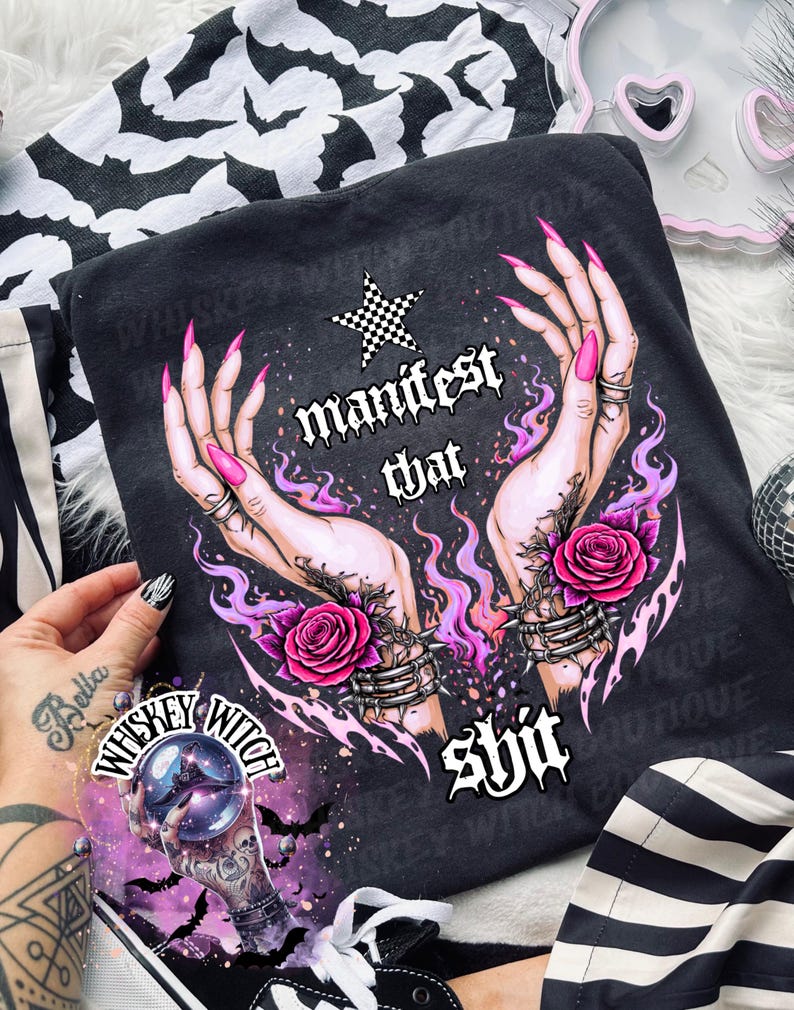 May include: Dark gray t-shirt with a graphic of hands, roses, and the text "manifest that shit." The design includes a checkered star and purple flames. A "Whiskey Witch" sticker is visible.