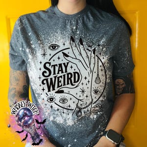 Handmade bleached styles shirt. Close-up of witchy Stay Weird shirt design with celestial artwork, mystical hands, stars, and bold black text