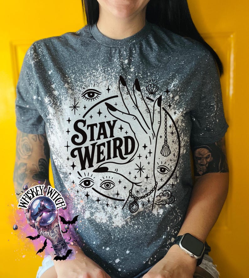 Handmade bleached styles shirt. Close-up of witchy Stay Weird shirt design with celestial artwork, mystical hands, stars, and bold black text