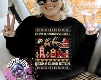 Può includere: Felpa nera con un design festivo con Babbo Natale su una slitta, renne e case. Il testo "SANTA KNOWS YOU'VE BEEN A DUMB BITCH" è visualizzato in un carattere pixelato. Parte della collezione Whiskey Witch Boutique.