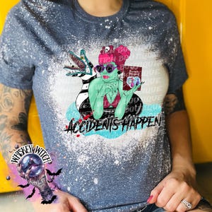 May include: A blue and white bleached t-shirt with a graphic of a woman with green skin and pink hair holding a book that says "Recently Deceased". The graphic also includes a green and black creature with a long tail. The text "Accidents Happen" is printed below the graphic.