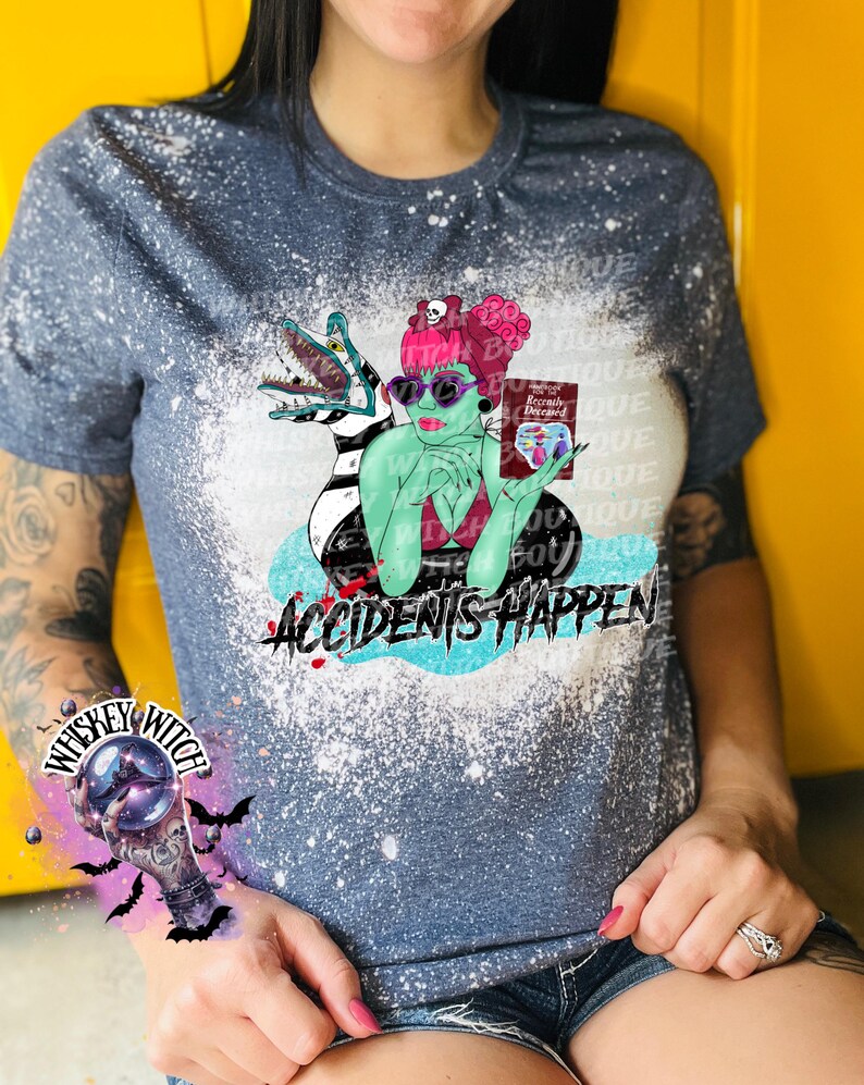 May include: A blue and white bleached t-shirt with a graphic of a woman with green skin and pink hair holding a book that says "Recently Deceased". The graphic also includes a green and black creature with a long tail. The text "Accidents Happen" is printed below the graphic.