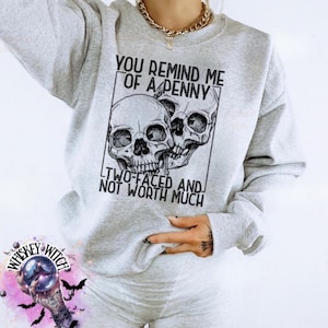 May include: A light gray sweatshirt with the text "YOU REMIND ME OF A PENNY TWO-FACED AND NOT WORTH MUCH" and a graphic of two skulls. The sweatshirt is part of a casual outfit.