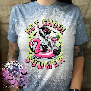 A indigo blue  and bleach splattered effect  t-shirt with a graphic of a zombie woman in a flamingo pool float, holding a cocktail. The text "Hot Ghoul Summer" is printed on the shirt.