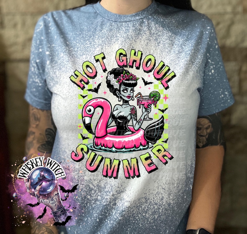 A indigo blue  and bleach splattered effect  t-shirt with a graphic of a zombie woman in a flamingo pool float, holding a cocktail. The text "Hot Ghoul Summer" is printed on the shirt.