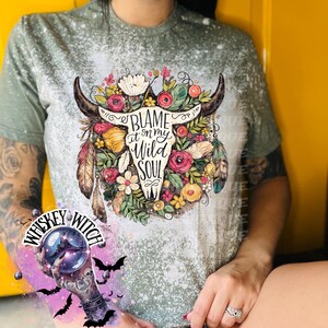 May include: A sage green t-shirt with a bleached design. The shirt features a graphic of a bull skull adorned with flowers and feathers, with the text "Blame it on my Wild Soul".