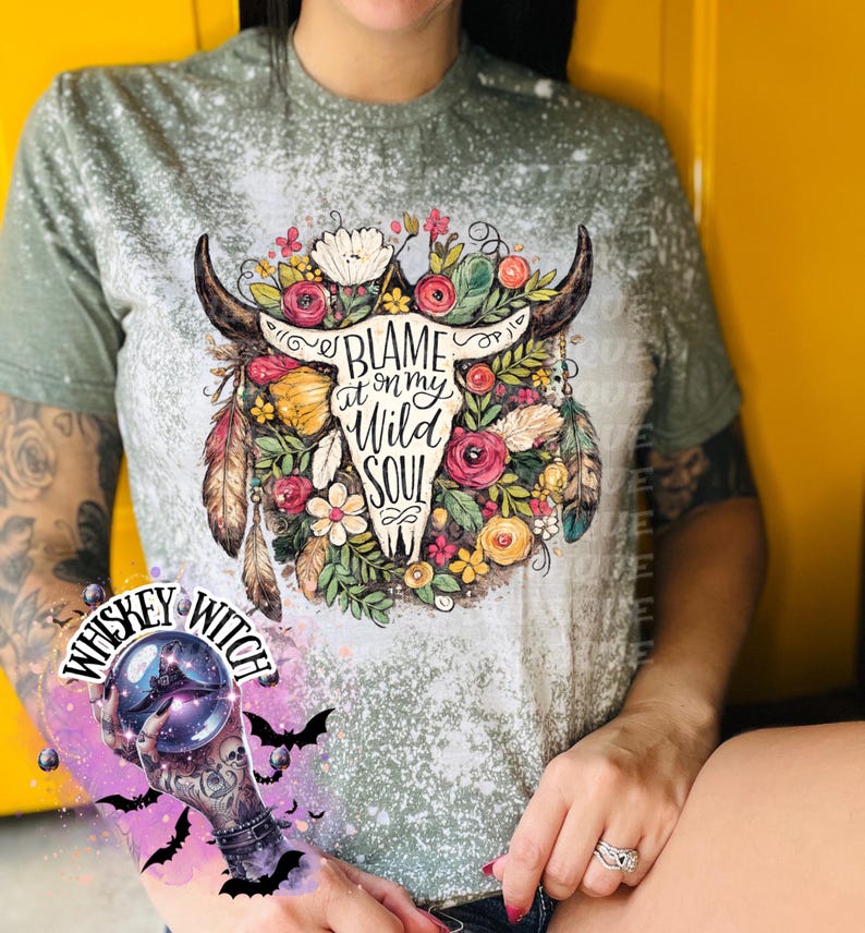 May include: A sage green t-shirt with a bleached design. The shirt features a graphic of a bull skull adorned with flowers and feathers, with the text "Blame it on my Wild Soul".