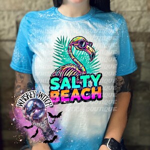May include: A blue and white tie-dye t-shirt featuring a skeleton flamingo wearing sunglasses, with the words "SALTY BEACH" in colorful text. The shirt has a crew neck and short sleeves.