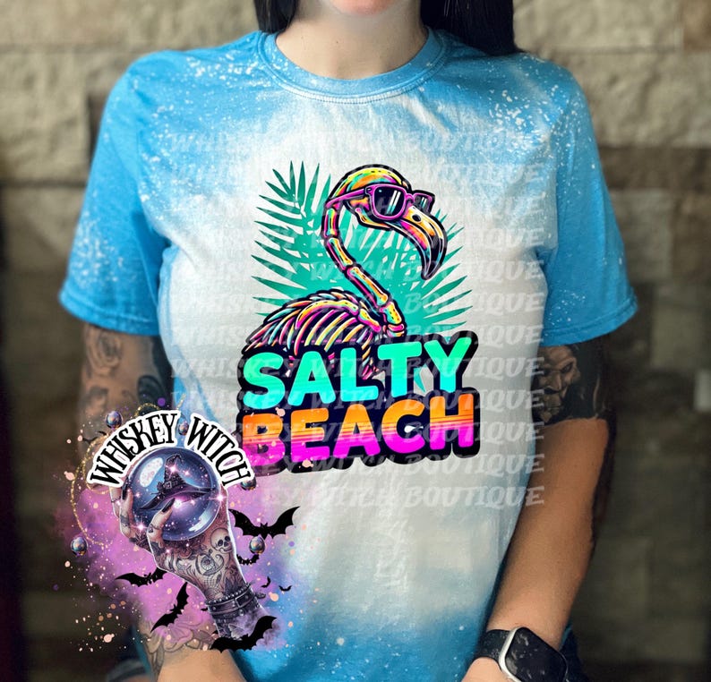 May include: A blue and white tie-dye t-shirt featuring a skeleton flamingo wearing sunglasses, with the words "SALTY BEACH" in colorful text. The shirt has a crew neck and short sleeves.