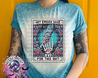 May include: A light blue t-shirt with a bleached design. The shirt features a graphic with the text "NOT ENOUGH SAGE FOR THIS SHIT". The graphic includes a skeleton hand holding a bundle of sage, surrounded by flowers.