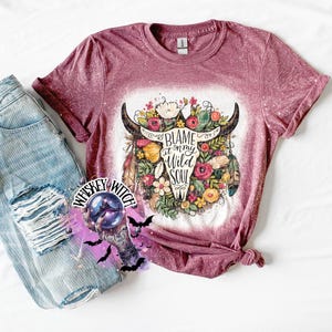 May include: A maroon t-shirt with a bleached design featuring a bull skull adorned with flowers and feathers. The text "Blame it on my Wild Soul" is printed on the skull. Light blue denim jeans are visible on the left.