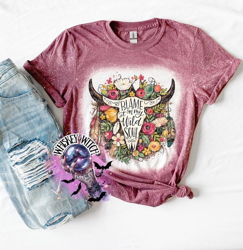May include: A maroon t-shirt with a bleached design featuring a bull skull adorned with flowers and feathers. The text "Blame it on my Wild Soul" is printed on the skull. Light blue denim jeans are visible on the left.