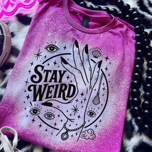 Handmade bleached styles shirt. Close-up of witchy Stay Weird shirt design with celestial artwork, mystical hands, stars, and bold black text