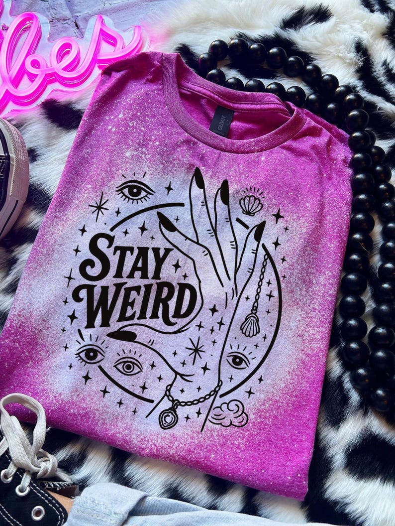 Handmade bleached styles shirt. Close-up of witchy Stay Weird shirt design with celestial artwork, mystical hands, stars, and bold black text