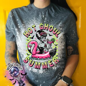 A grey and bleach splattered effect  t-shirt with a graphic of a zombie woman in a flamingo pool float, holding a cocktail. The text "Hot Ghoul Summer" is printed on the shirt.