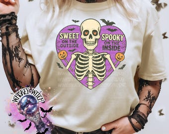 May include: A cream-coloured t-shirt with a purple heart design featuring a skeleton graphic. The heart reads "Sweet on the Outside, Spooky on the Inside." The skeleton holds pumpkins. Bats and "Whiskey Witch" text are also visible.