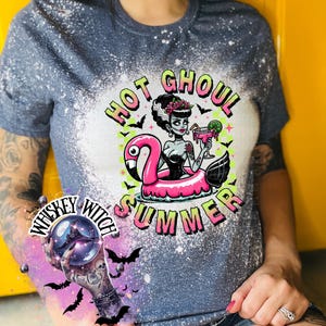A navy blue  and bleach splattered effect  t-shirt with a graphic of a zombie woman in a flamingo pool float, holding a cocktail. The text "Hot Ghoul Summer" is printed on the shirt.