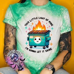 green t-shirt with a bleach-splattered design featuring a cartoon dumpster on fire with the text "This little light of mine I'm gonna let it shine".