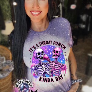 May include: Purple t-shirt with a graphic of two skeletons fist bumping. The skeletons are wearing boxing gloves and the text reads "It's a throat punch kinda day".