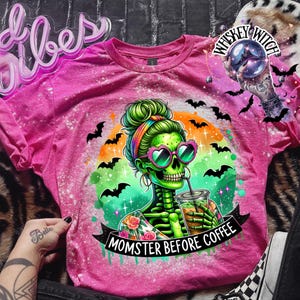 May include: A pink t-shirt with a graphic of a green skeleton woman wearing heart-shaped sunglasses, drinking from a cup. The shirt has the text "MOMSTER BEFORE COFFEE" and is decorated with bats and a rainbow. The shirt has a bleached effect.