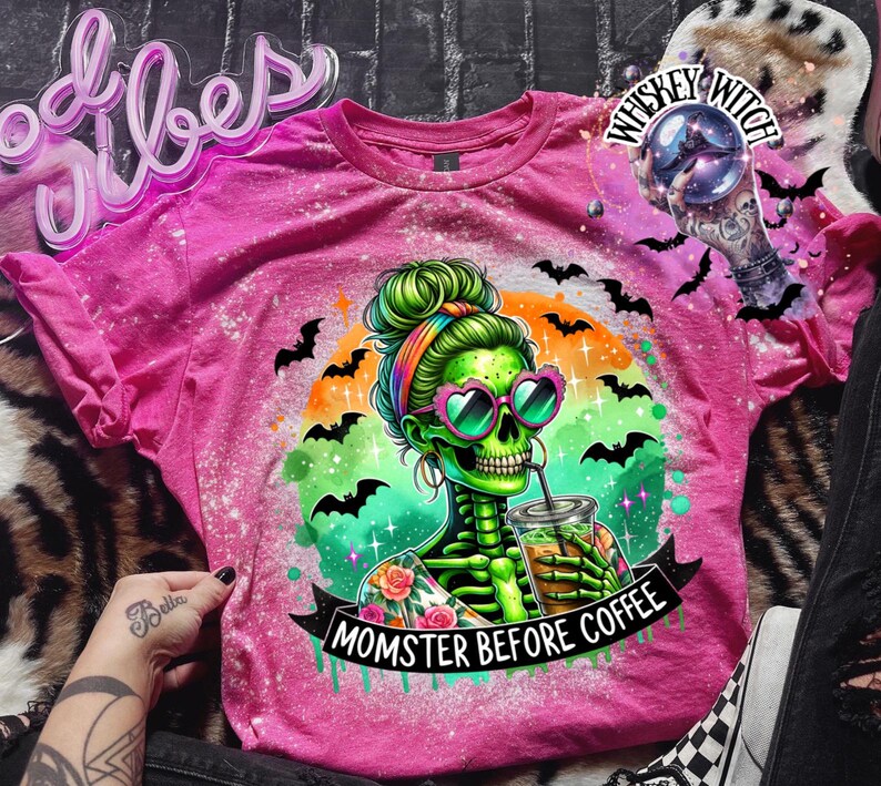 May include: A pink t-shirt with a graphic of a green skeleton woman wearing heart-shaped sunglasses, drinking from a cup. The shirt has the text "MOMSTER BEFORE COFFEE" and is decorated with bats and a rainbow. The shirt has a bleached effect.