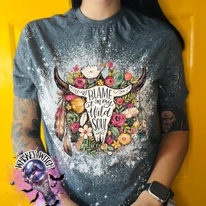 May include: A gray t-shirt with a bleached design featuring a bull skull adorned with flowers and feathers. The text "BLAME it on my WILD SOUL" is centered on the skull. The shirt has a crew neck and short sleeves.