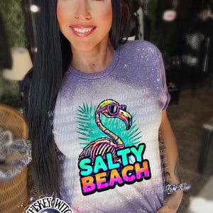 May include: A purple t-shirt with a flamingo graphic wearing sunglasses and the words "Salty Beach". The shirt has a bleached effect and is worn by a person. The image also includes the text "Whiskey Witch".