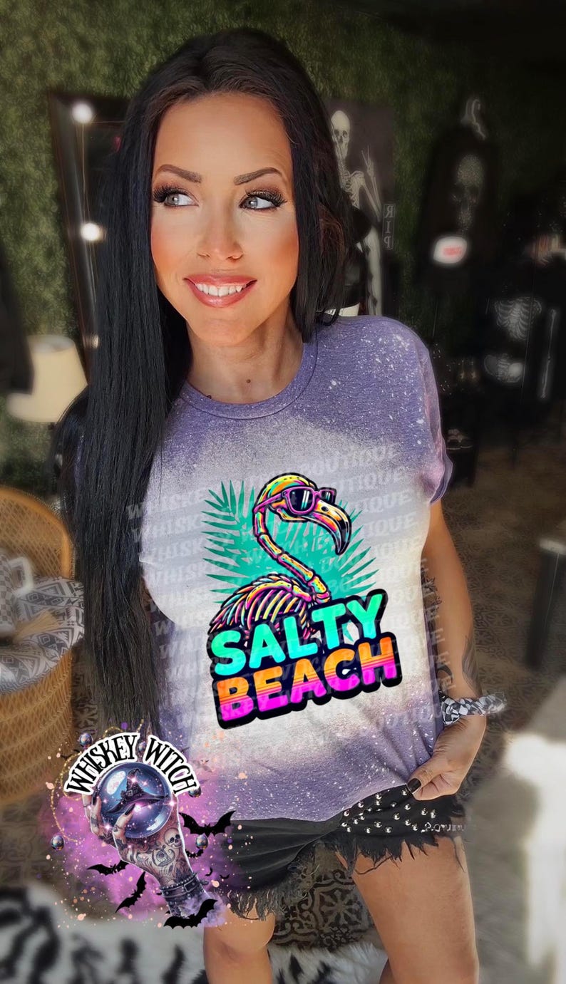 May include: A purple t-shirt with a flamingo graphic wearing sunglasses and the words "Salty Beach". The shirt has a bleached effect and is worn by a person. The image also includes the text "Whiskey Witch".