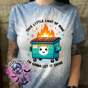 A light blue t-shirt with a bleach-splattered design. The shirt features a cartoon image of a smiling trash can with flames coming out of the top. The text "This little light of mine I'm gonna let it shine" is printed on the shirt.